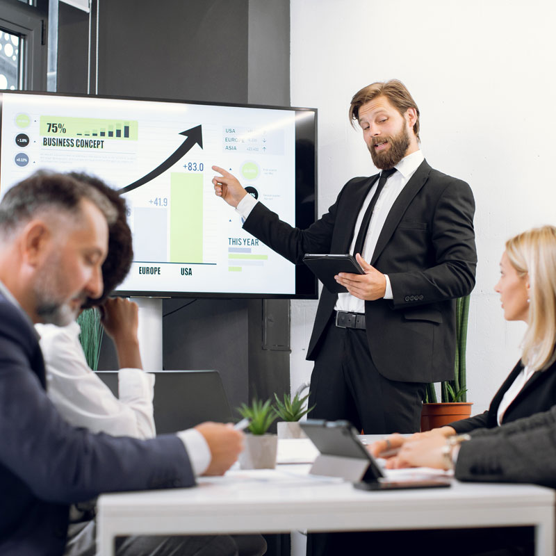 Business presentation with a speaker in a suit pointing at a growth chart on a screen, illustrating strategic growth metrics in a corporate meeting setting.