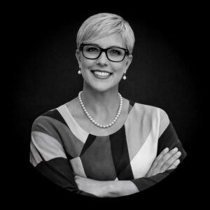Professional woman with short blonde hair and glasses, wearing a colorful patterned blouse and pearl necklace, smiling confidently against a dark background, representing leadership and commitment in consulting for mission-driven organizations.